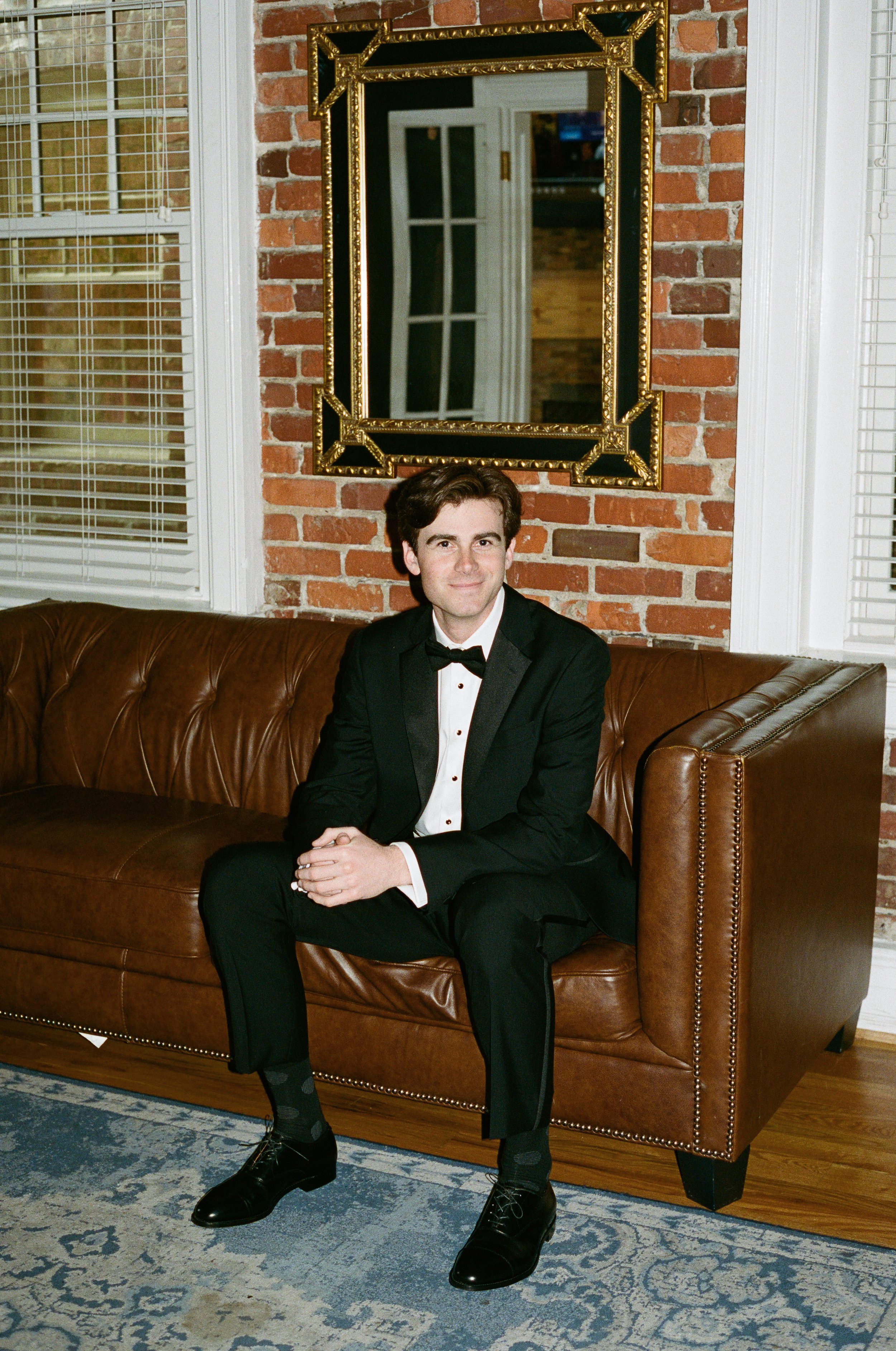 A man in a tuxedo sitting on a brown leather couch in front of a brick wall with a gold and black framed mirror, and a window with white blinds. richmond virginia wedding photographer