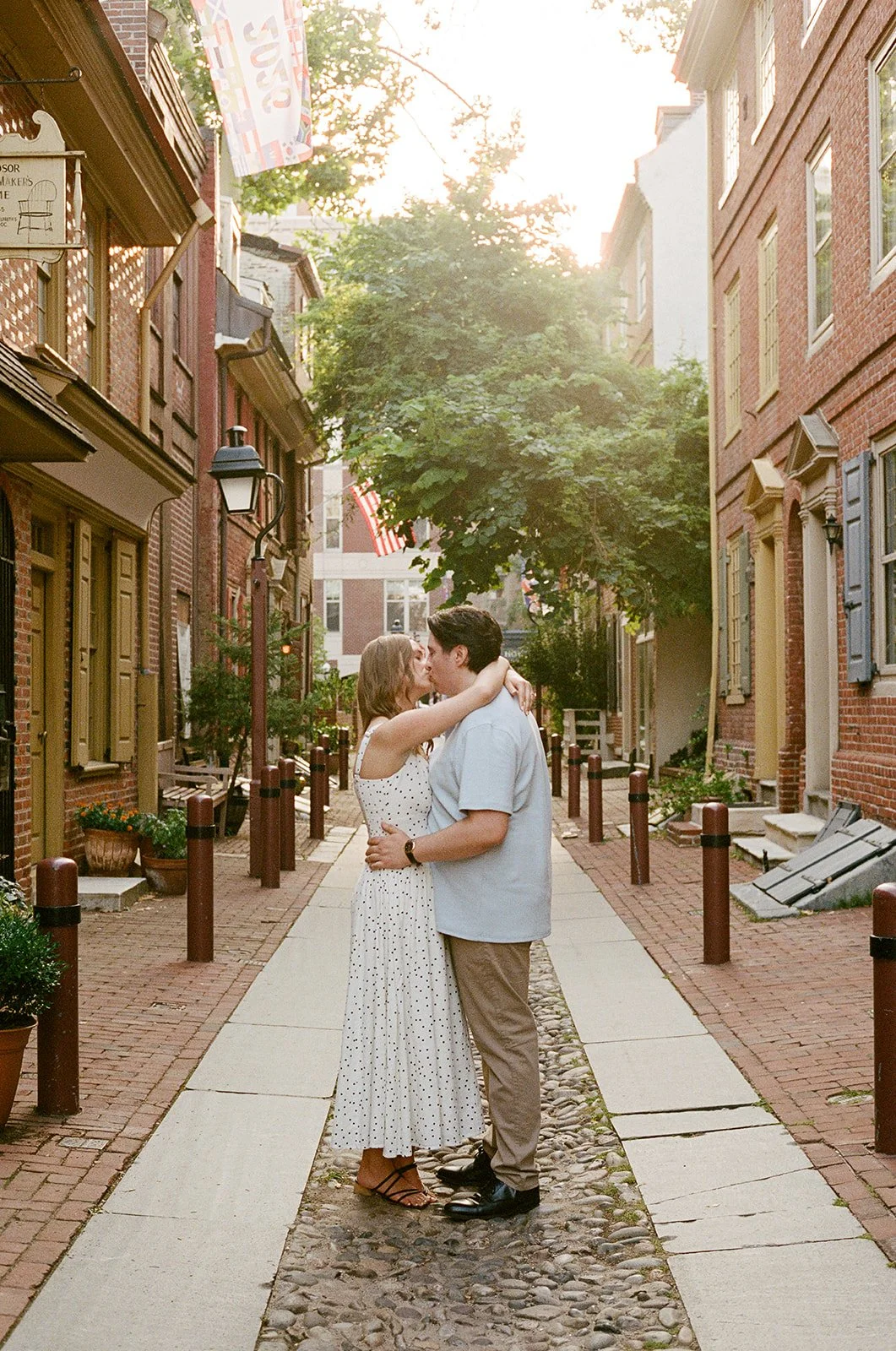 A couple sharing a kiss and embracing in a quaint brick alleyway during sunset, surrounded by colorful buildings and greenery. Old City Philadelphia engagement shoot