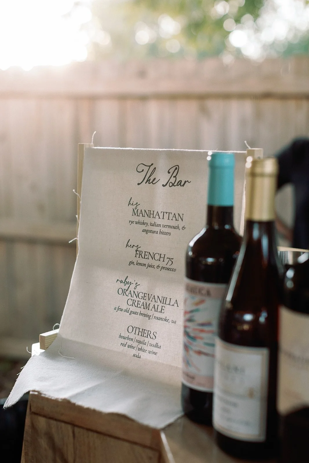 A menu titled 'The Bar' listing drinks including Manhattan, French 75, Orange Vanilla Cream Ale, and other options, with various bottles of alcohol in the foreground and a wooden fence in the background. Roanoke Virginia Wedding Film Photographer.