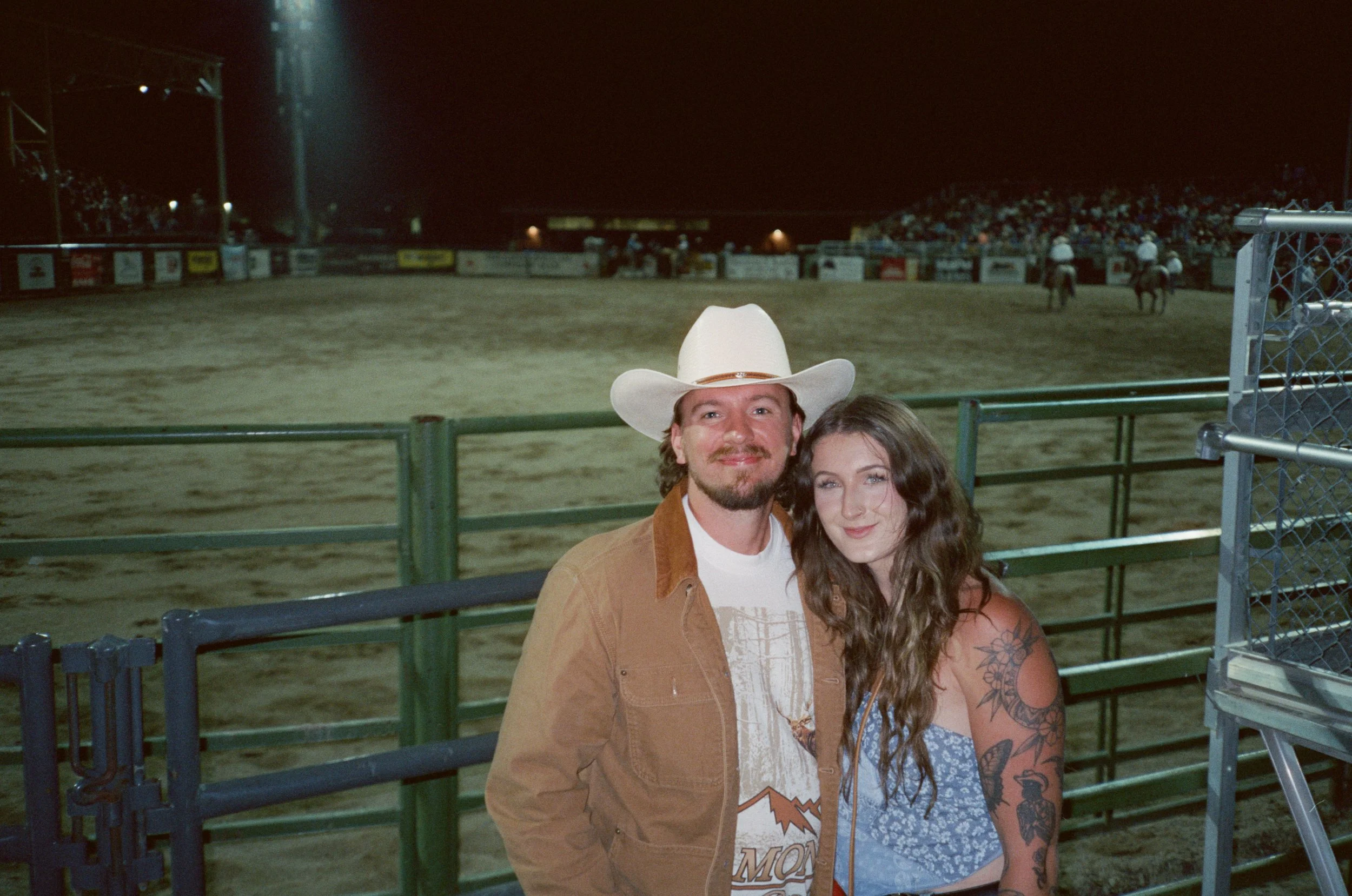 A man and woman stand together at a rodeo arena at night, smiling. The man wears a white cowboy hat and a brown jacket, and the woman has long wavy hair with tattoos on her arm, wearing a blue patterned top. Jackson Wyoming.