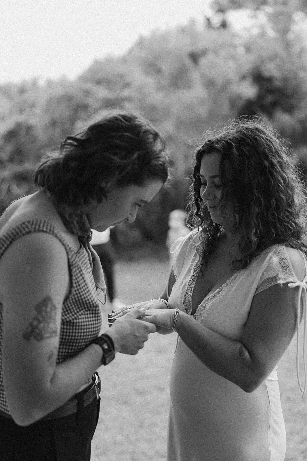 Two women, one with curly hair and the other with wavy hair, standing outdoors, holding hands and smiling, with a blurred natural background. ocracoke outerbanks wedding film photographer