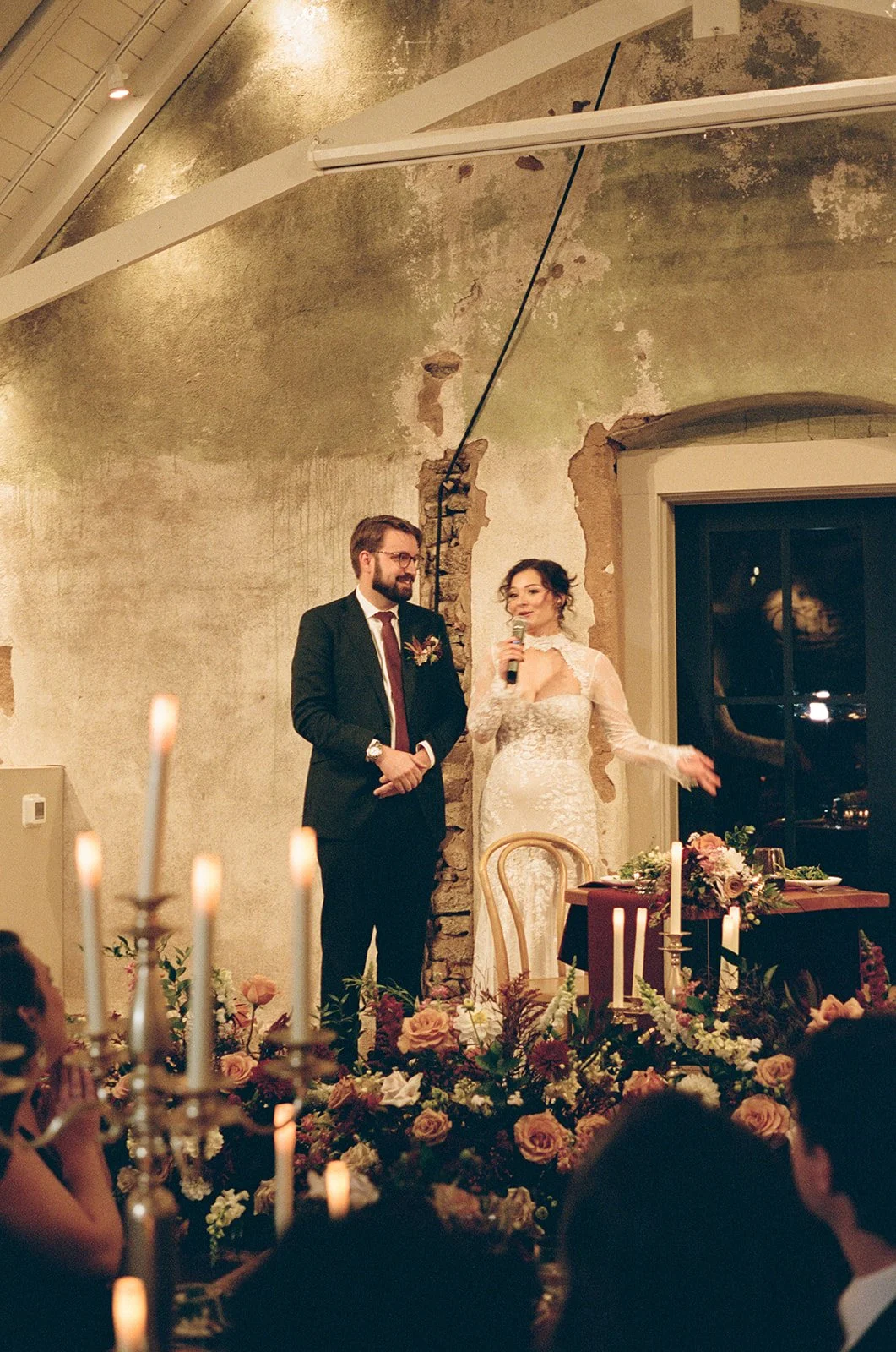 A bride in a white lace wedding dress speaking into a microphone at her wedding reception, standing next to the groom in a dark suit, in front of a rustic decorated table with floral arrangements and candles. potters craft cider charlottesville va