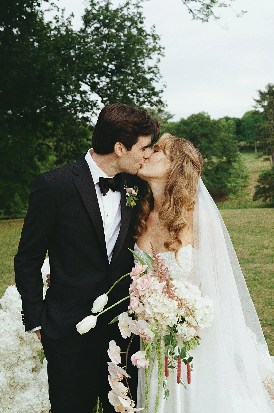 A newlywed couple kissing outdoors, with the groom in a black tuxedo and bow tie, and the bride in a white wedding dress holding a large bouquet of pink, white, and green flowers.  richmond wedding film photographer virginia
