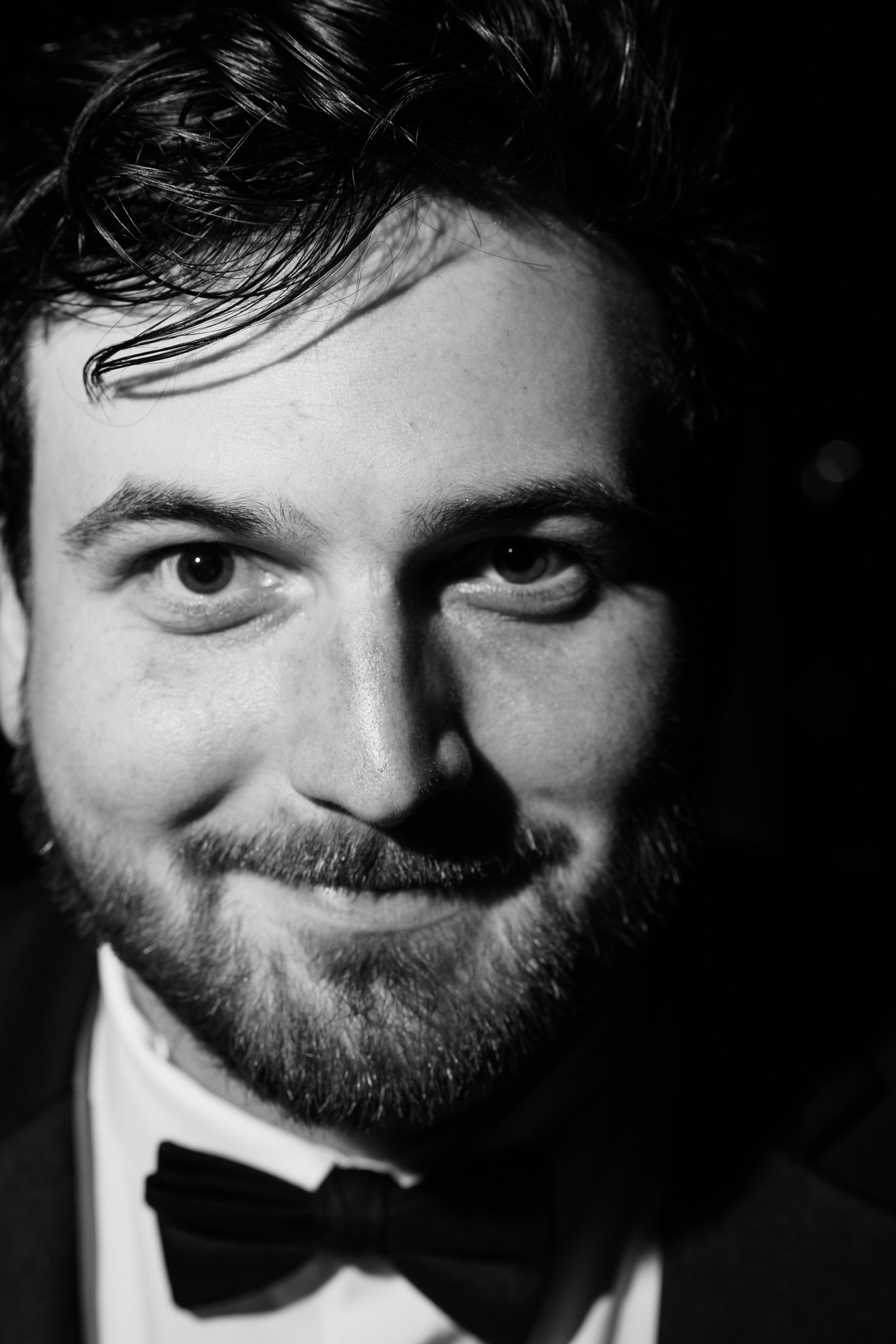 Close-up black and white photo of a man's face, smiling, wearing a tuxedo with a bow tie, with dark curly hair.