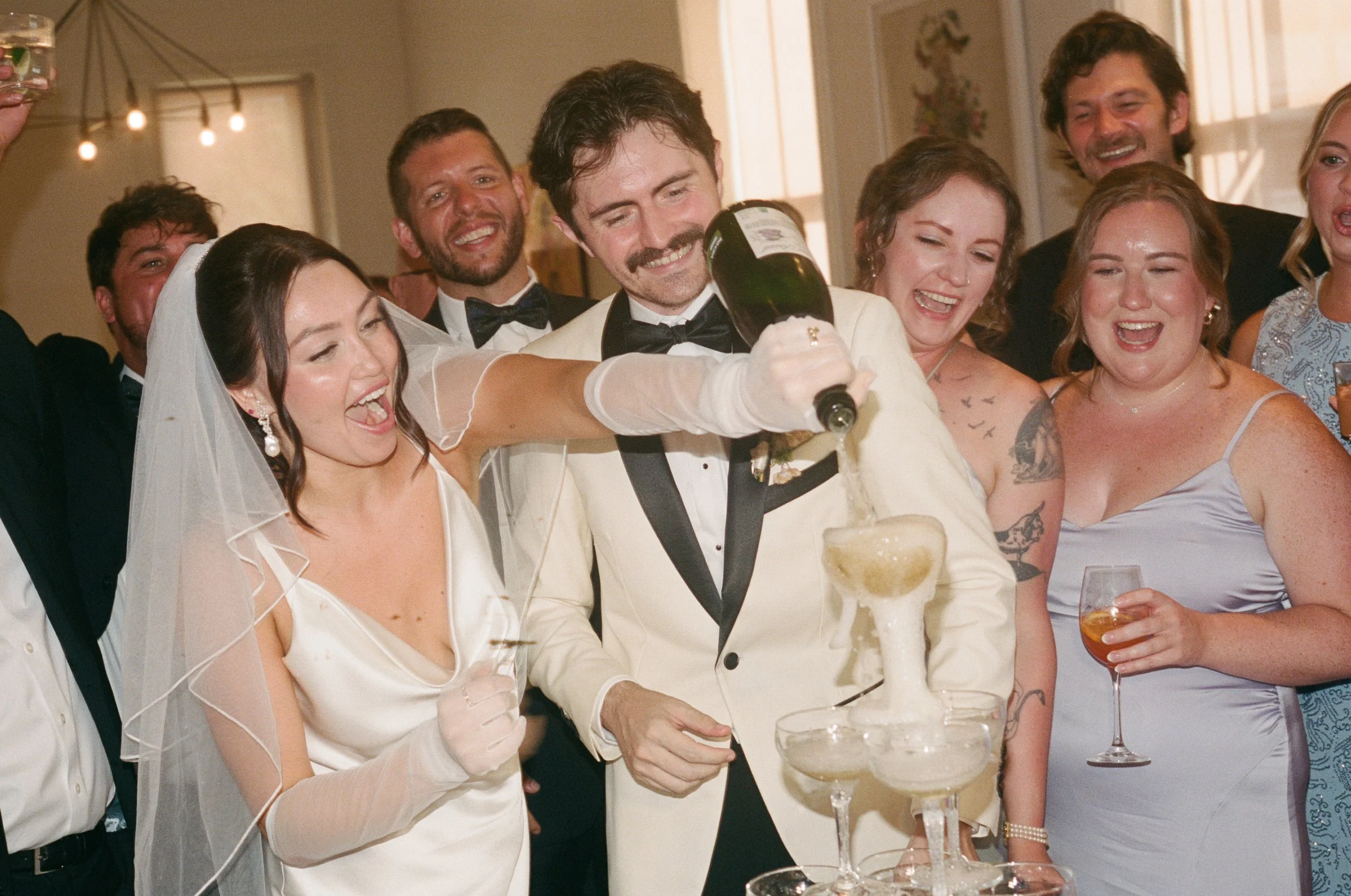 Group of wedding guests celebrating, with a woman in a veil pouring champagne into a tower of glasses, surrounded by smiling friends in formal attire at Common House in Richmond Virginia.