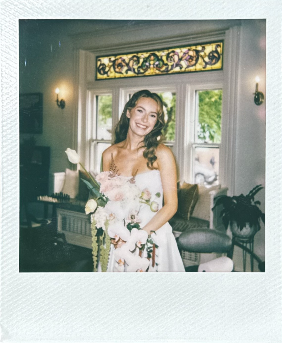 A young woman in a wedding dress holding a bouquet of flowers standing in a room with large windows and stained glass decor, smiling.