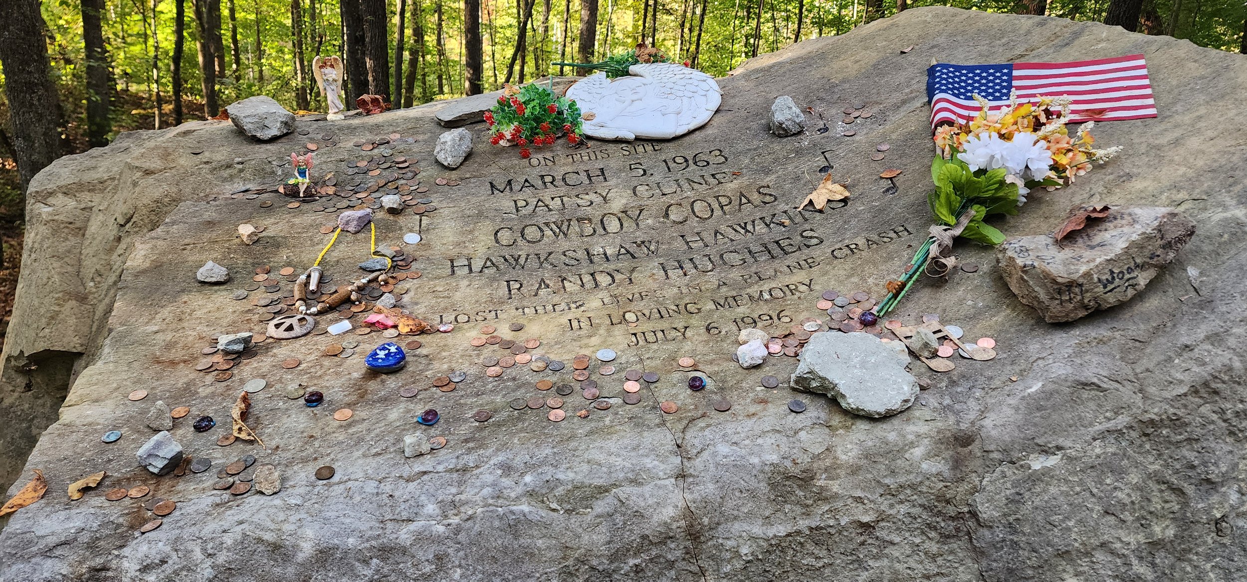 A large rock with trinkets, flowers and a flag surrounding the information about the crash that killed Patsy Cline