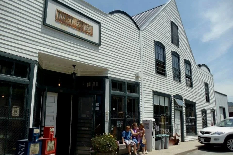An old wooden building with an old gas pump and two newspaper machines with a rusty sign above the door saying Mast General Store