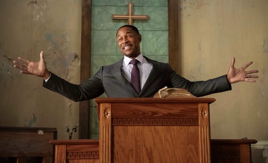 A pastor in a gray suit stands behind a wooden pulpit with his arms spread wide in a church with peeling walls and a cross behind him.