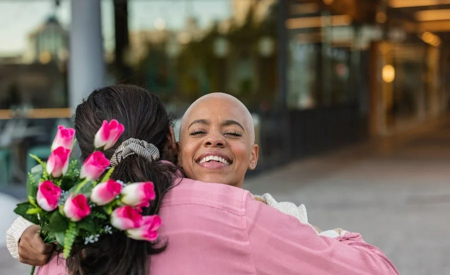 A smiling woman with a shaved head hugs a friend who is holding a bouquet of pink roses.