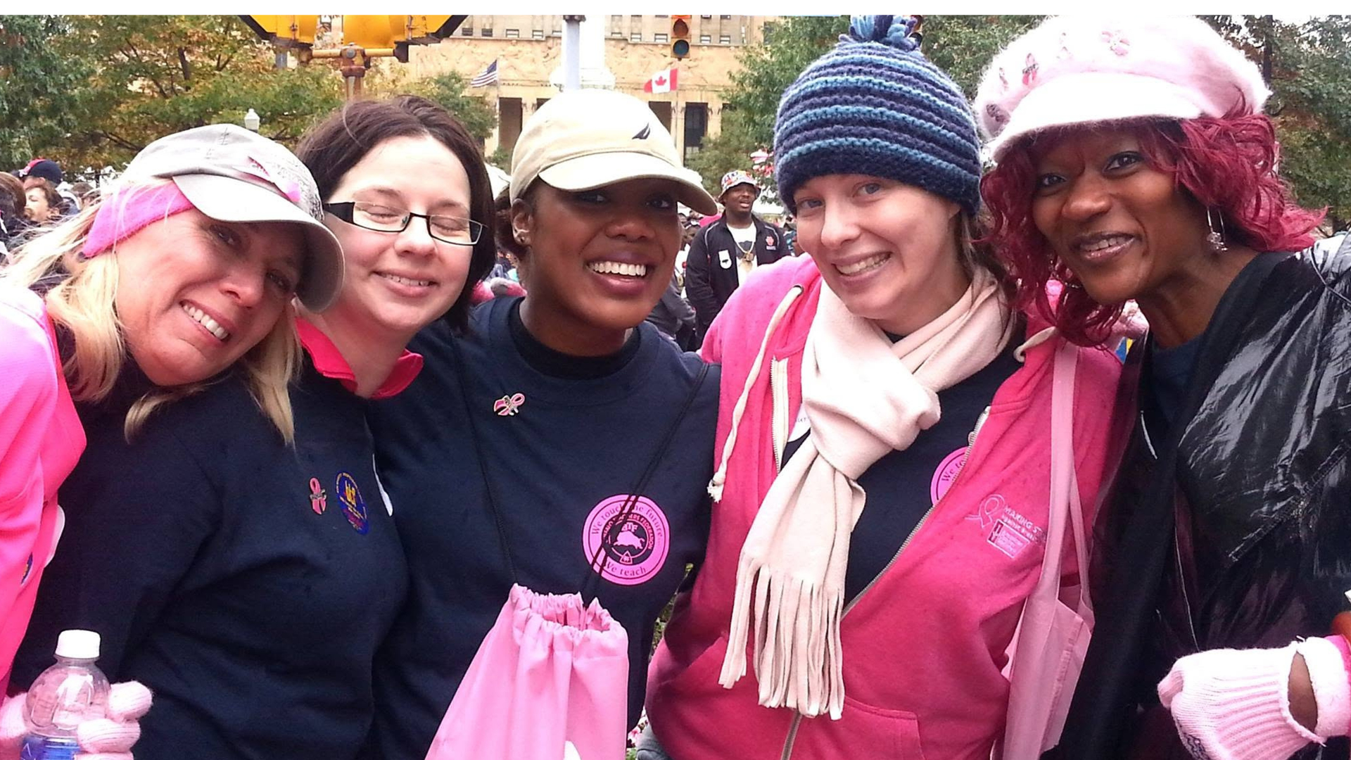 Five smiling women in pink and navy breast cancer awareness gear pose together outdoors at a downtown walk, with Dianna Hobbs in the center wearing a tan baseball cap.