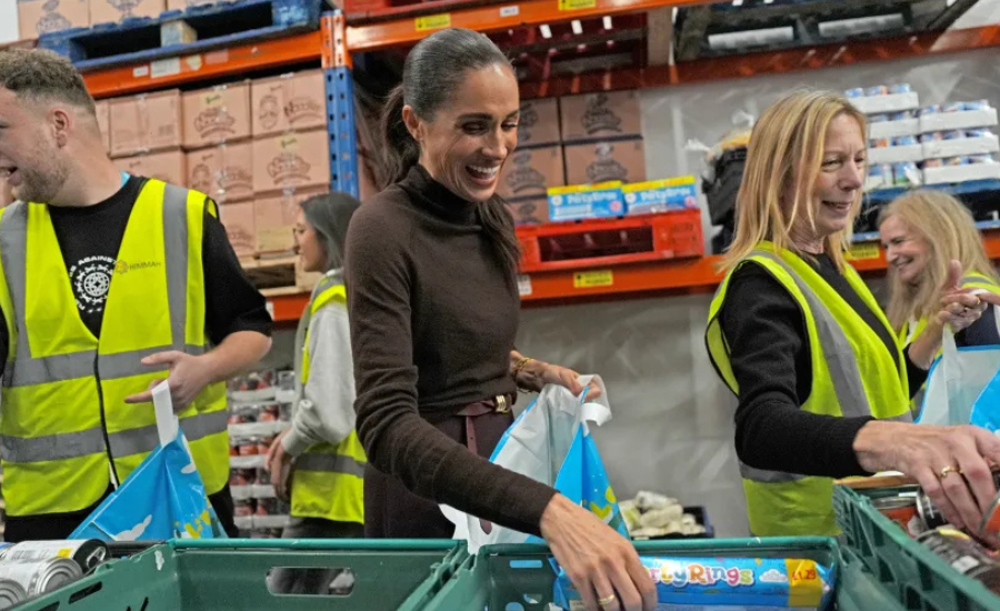 Meghan, smiling, packs groceries into blue bags at a food bank warehouse alongside volunteers in yellow high-visibility vests, with shelves of boxed food behind them.