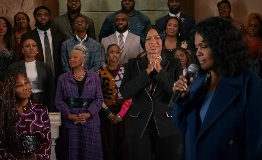 A congregation stands in a church as a woman in a blue coat sings into a microphone and a woman in black at center clasps her hands in prayer.