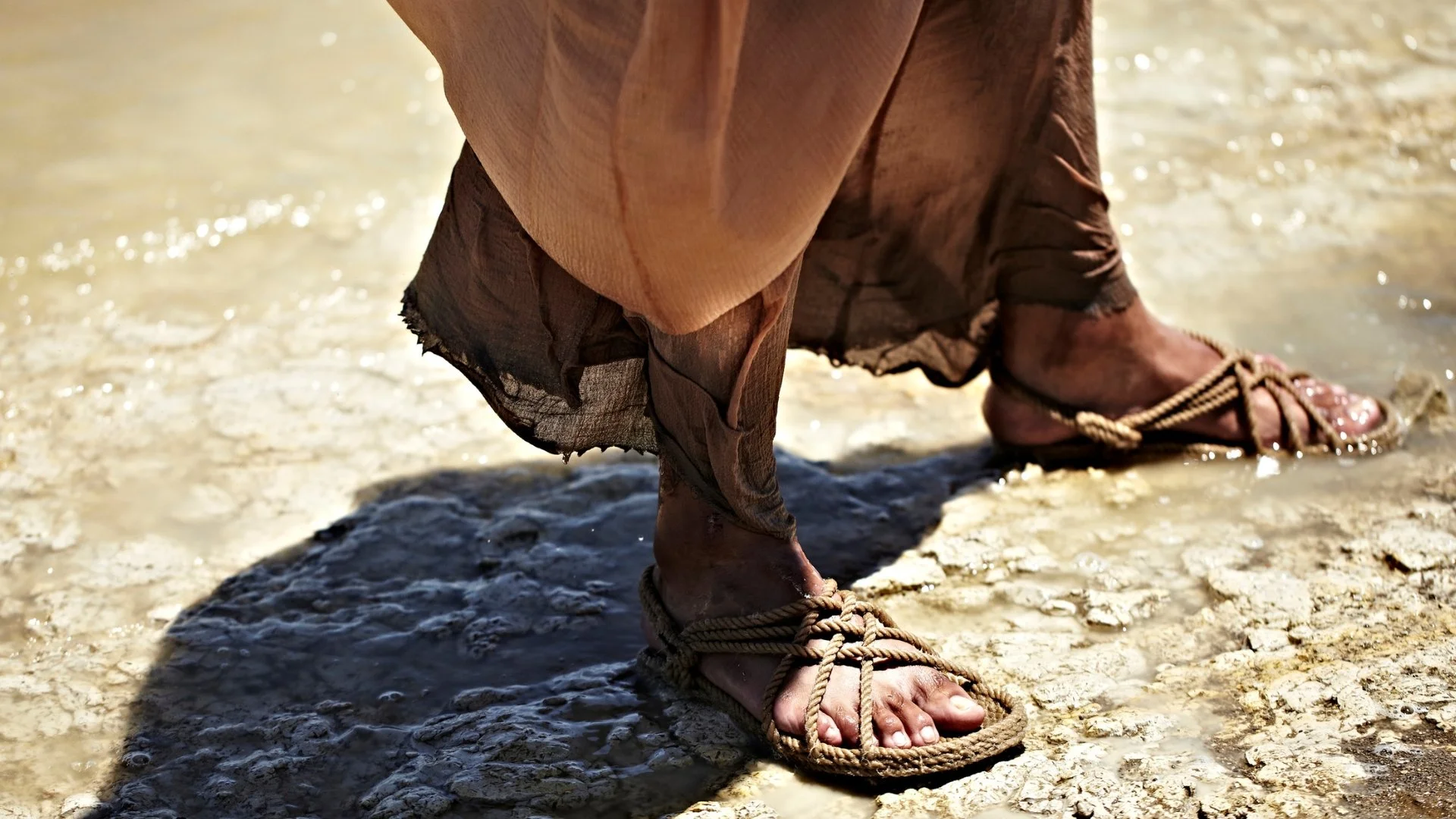 Close-up of feet in rope sandals beneath the frayed hem of a linen robe, standing on wet, sunlit ground.