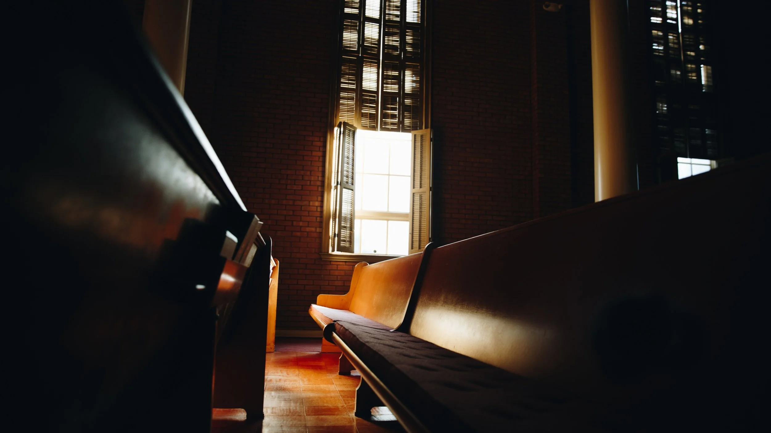Sunlight streams through an open shuttered window into an empty church sanctuary lined with wooden pews.