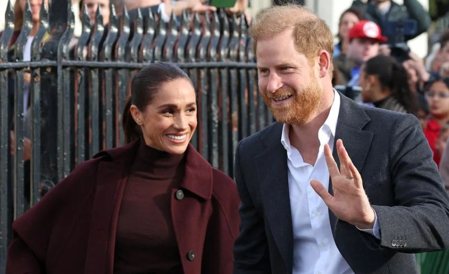 Harry waves and smiles beside Meghan in front of a black iron fence, with onlookers behind them.