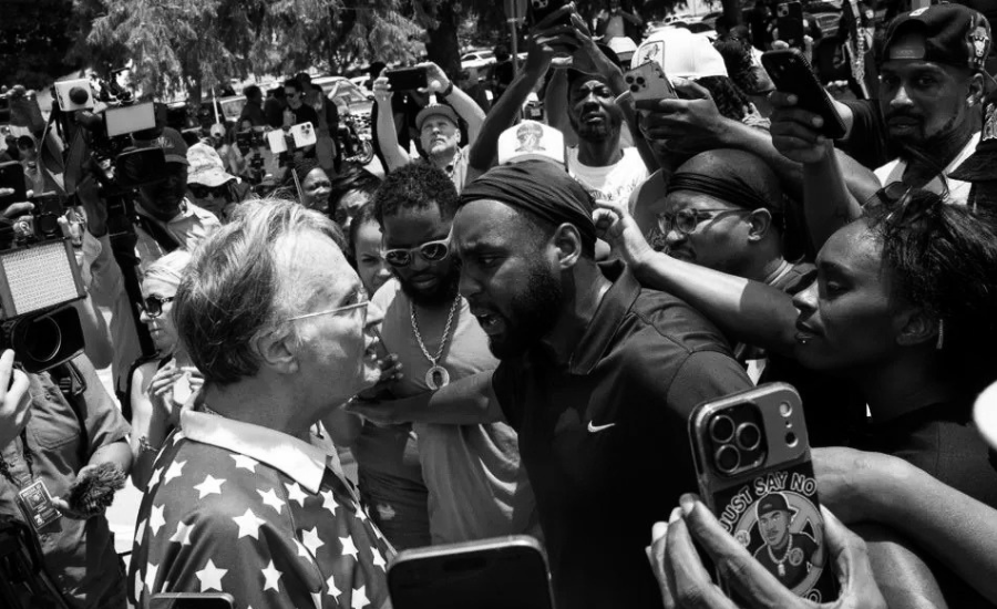 Black and white photo of a man in a star-patterned shirt and a younger man in a dark polo shirt and headwrap standing face to face in apparent confrontation, surrounded by a crowd holding up phones and cameras to record the scene.