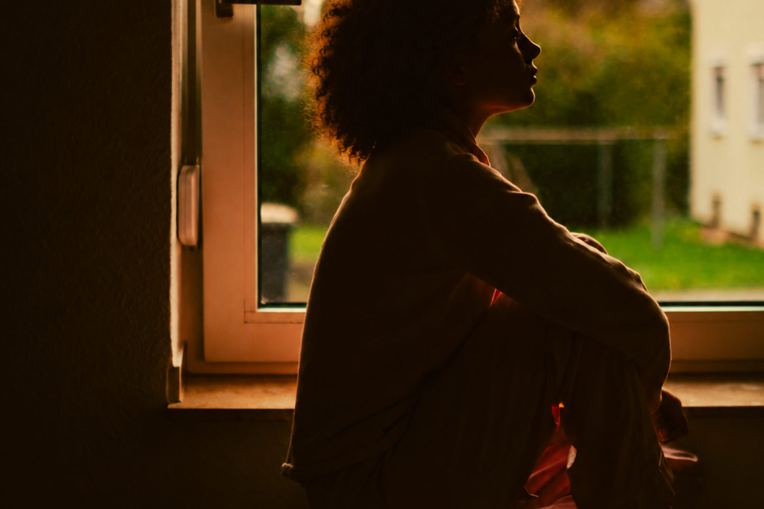 A woman with curly hair sits in silhouette beside a window, knees drawn up, gazing out toward a green yard in warm afternoon light.