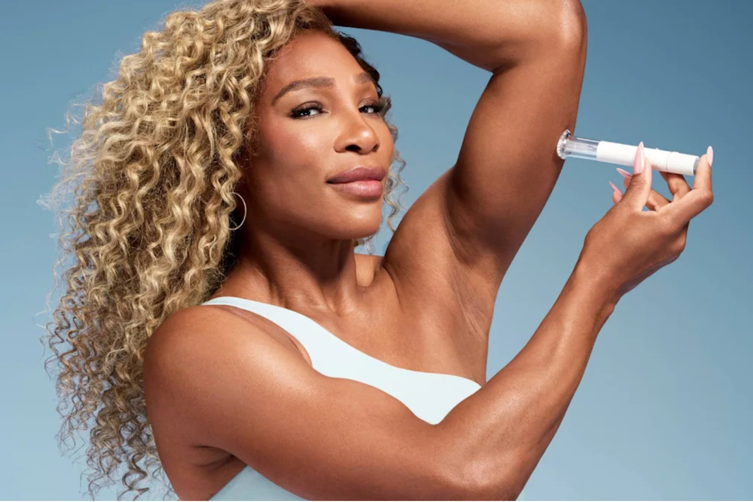 Williams, with long curly blonde hair and a white top, holds an injection pen to her upper arm against a blue background.