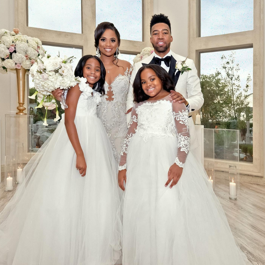 LaShonda Moore and Marlon Moore pose with two young girls in white gowns on their wedding day at an elegant venue with floor-to-ceiling windows and floral arrangements