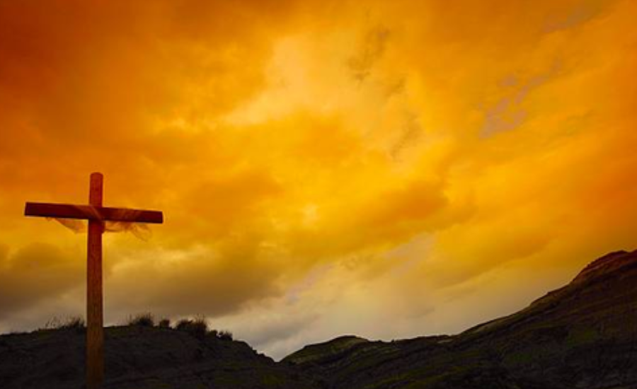 Wooden cross silhouetted against a vivid orange and gold sunset sky over dark hills