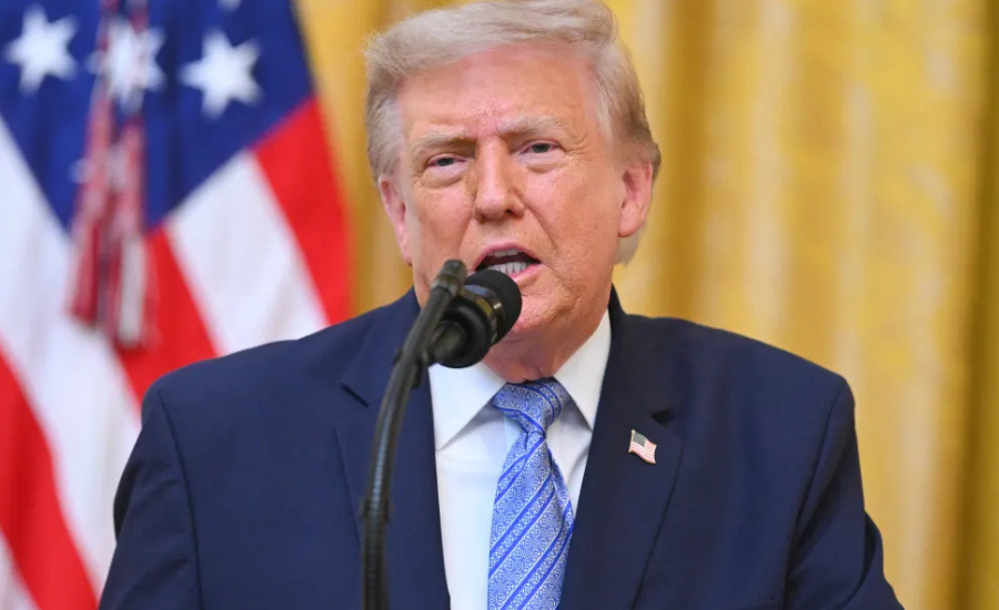 President Trump speaking at a podium in front of American flags and a gold curtain backdrop.