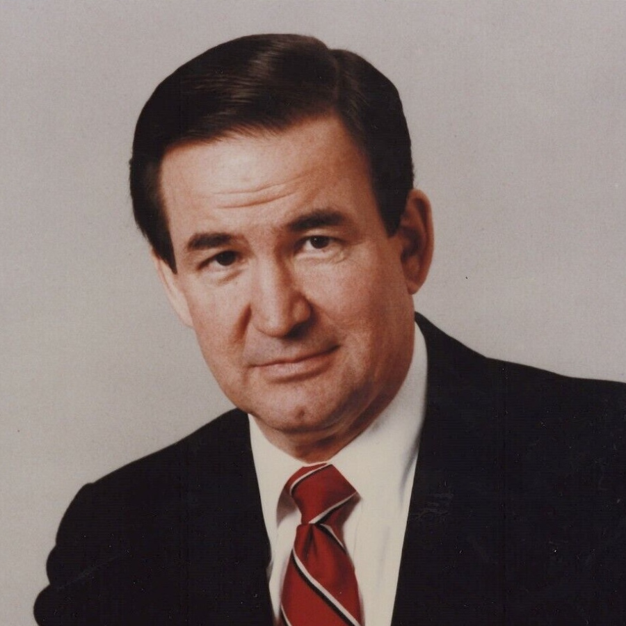 Formal portrait of Pat Buchanan wearing a dark suit, white shirt and red striped tie against a plain light background.