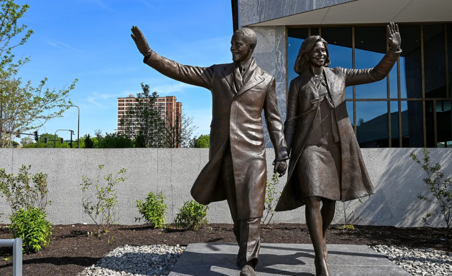 Bronze statue of Barack and Michelle Obama waving, standing outside the Obama Presidential Center in Chicago with landscaping and a residential building visible in the background