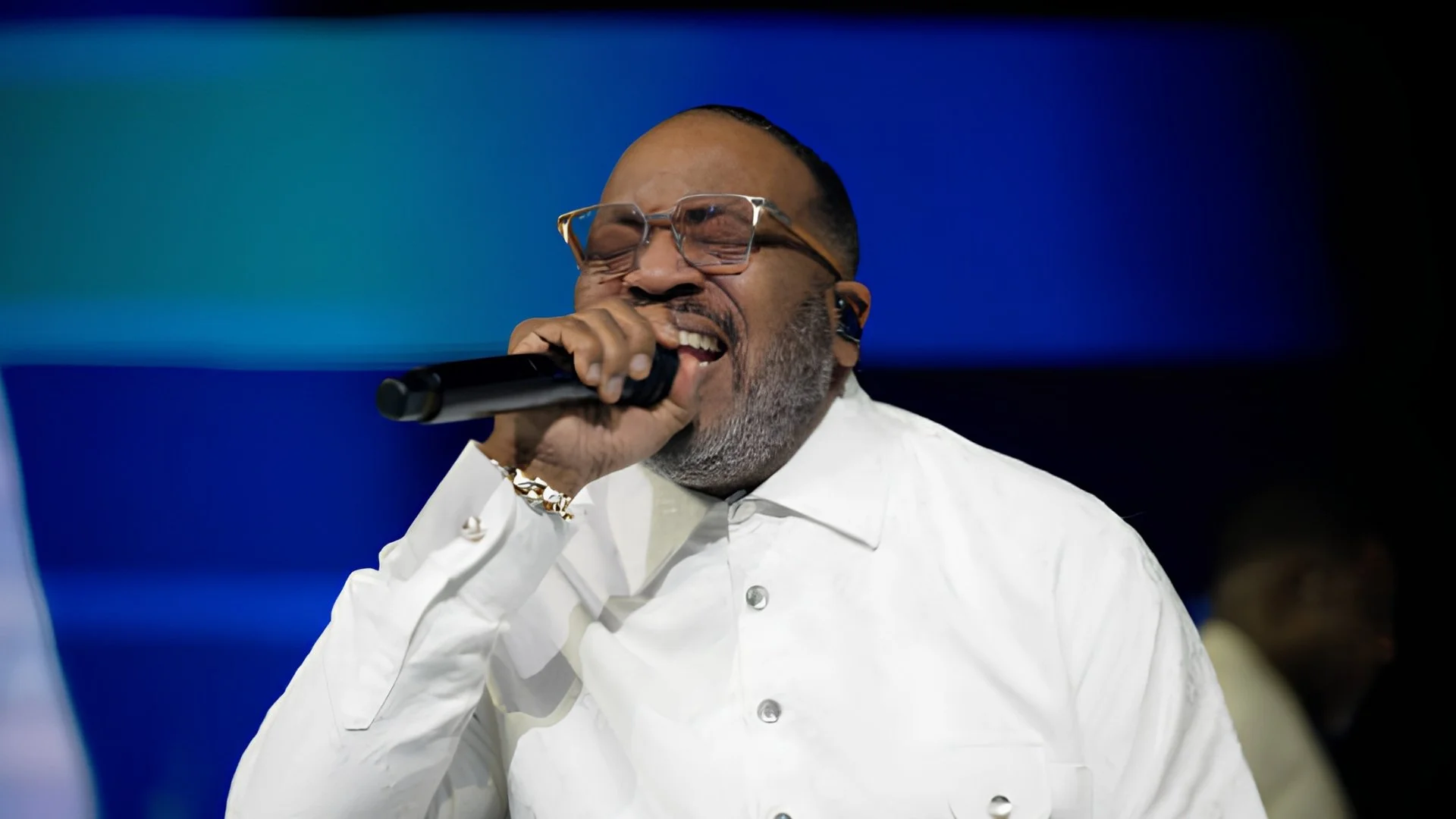 Marvin Sapp, wearing a white shirt and clear-framed glasses, sings into a microphone with his eyes closed in front of blue stage lights.
