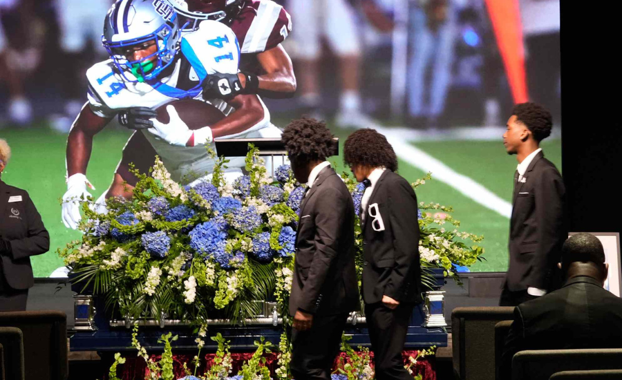 Young men in dark suits pass a flower-covered casket while football game footage plays on a large screen behind them