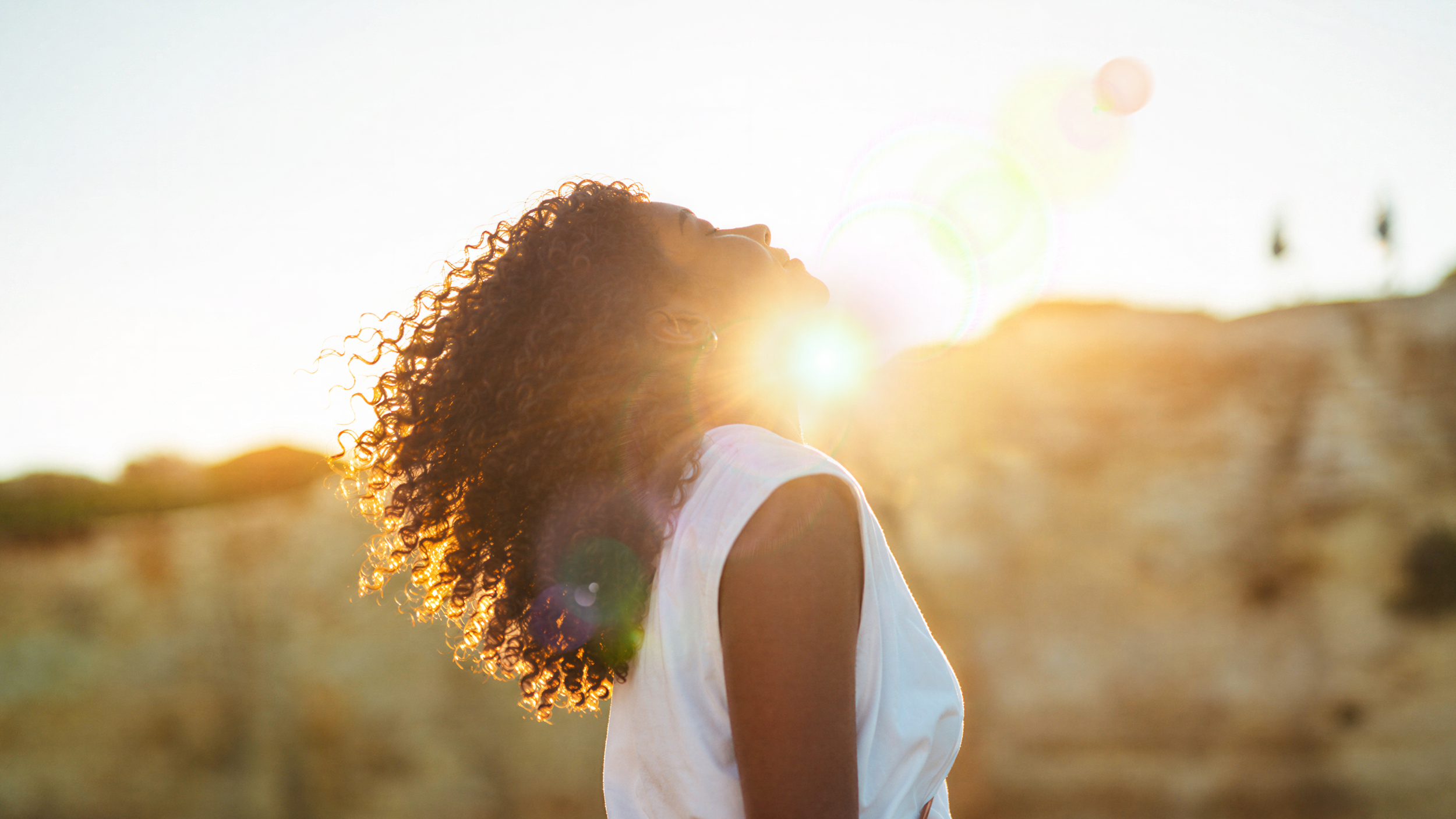 A woman with curly hair tilts her head back with her eyes closed as golden sunlight shines around her.