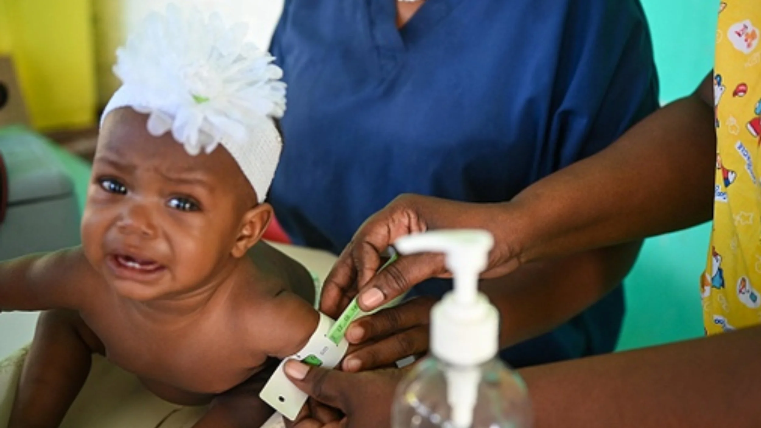 A crying baby in a white flower headband has a green-marked measuring tape wrapped around her upper arm by a health worker in blue scrubs.