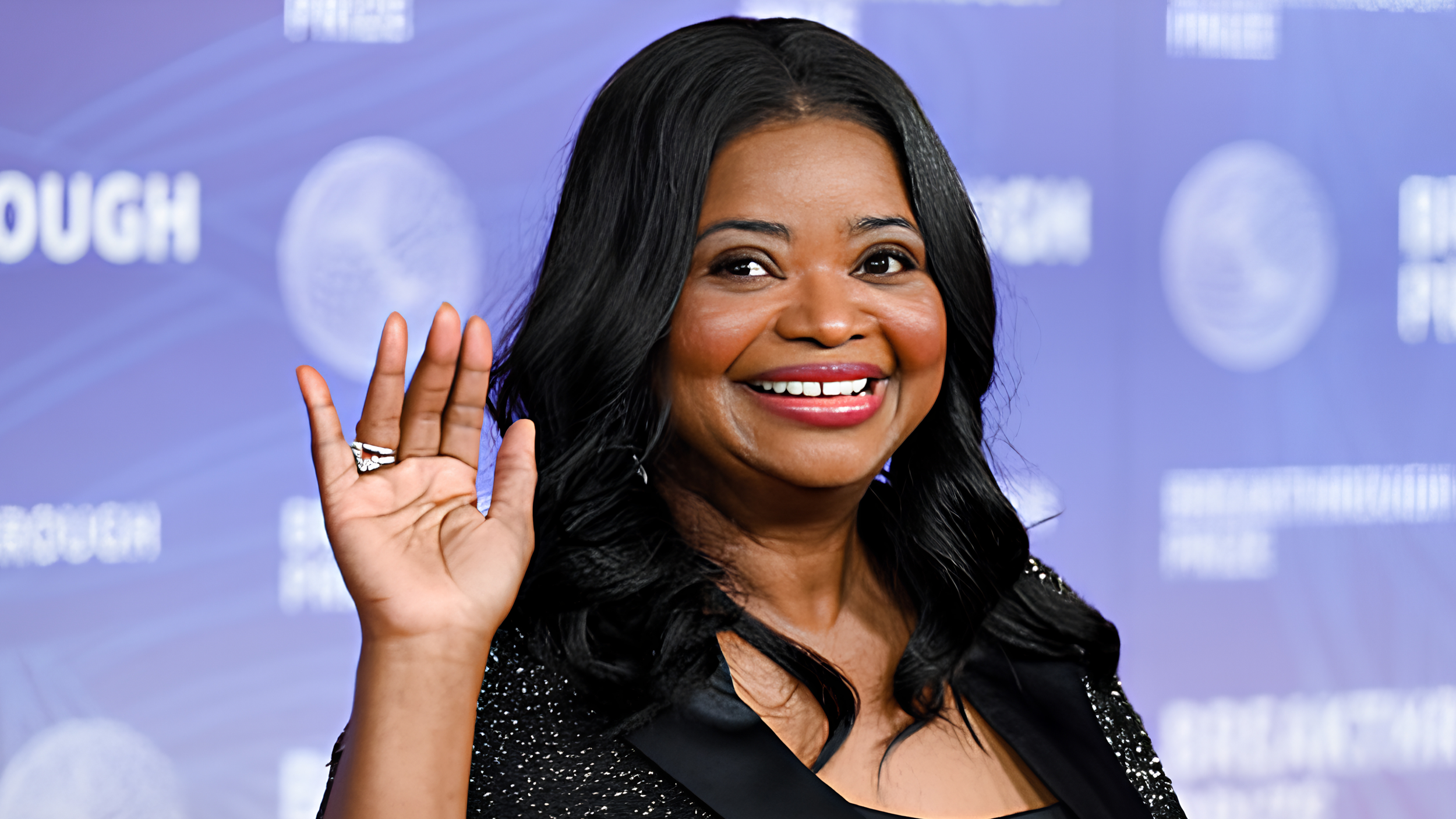Octavia Spencer smiles and waves on the red carpet at the Breakthrough Prize Ceremony, wearing a black sequined jacket with satin lapels, in front of a lavender event backdrop.