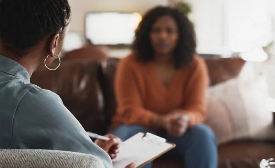 A woman holding a clipboard listens to a client seated across from her on a couch during a counseling session.