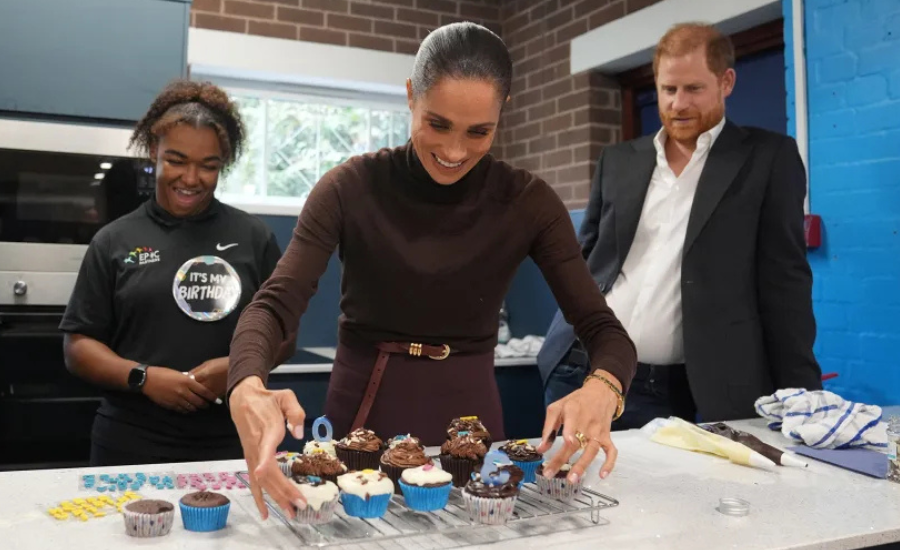 Meghan, smiling, arranges frosted cupcakes on a cooling rack in a kitchen as a young woman wearing an Epic Partners shirt and an "It's my birthday" badge looks on, with Harry standing behind them.