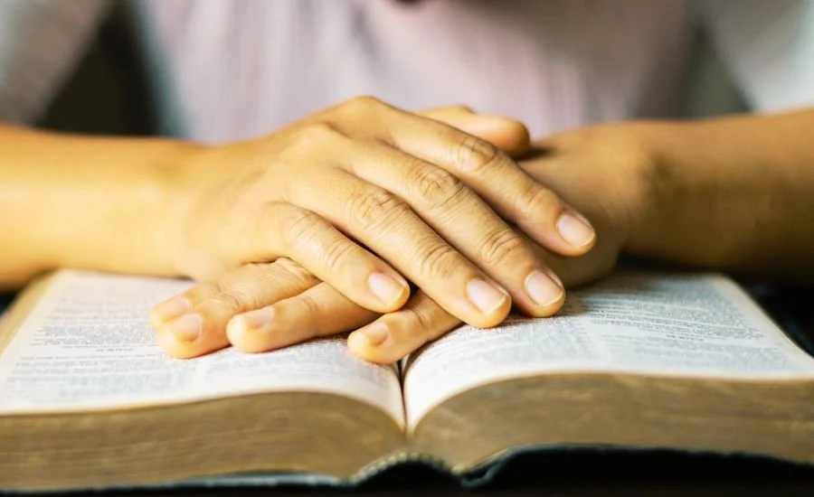 A person’s folded hands rest on the pages of an open Bible in a posture of prayer.