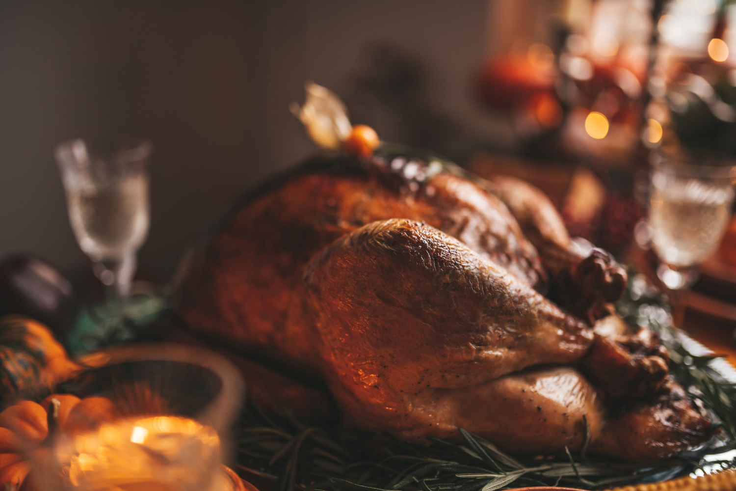 A golden roast turkey on a bed of rosemary, surrounded by candlelight, glassware and small pumpkins on a Thanksgiving table