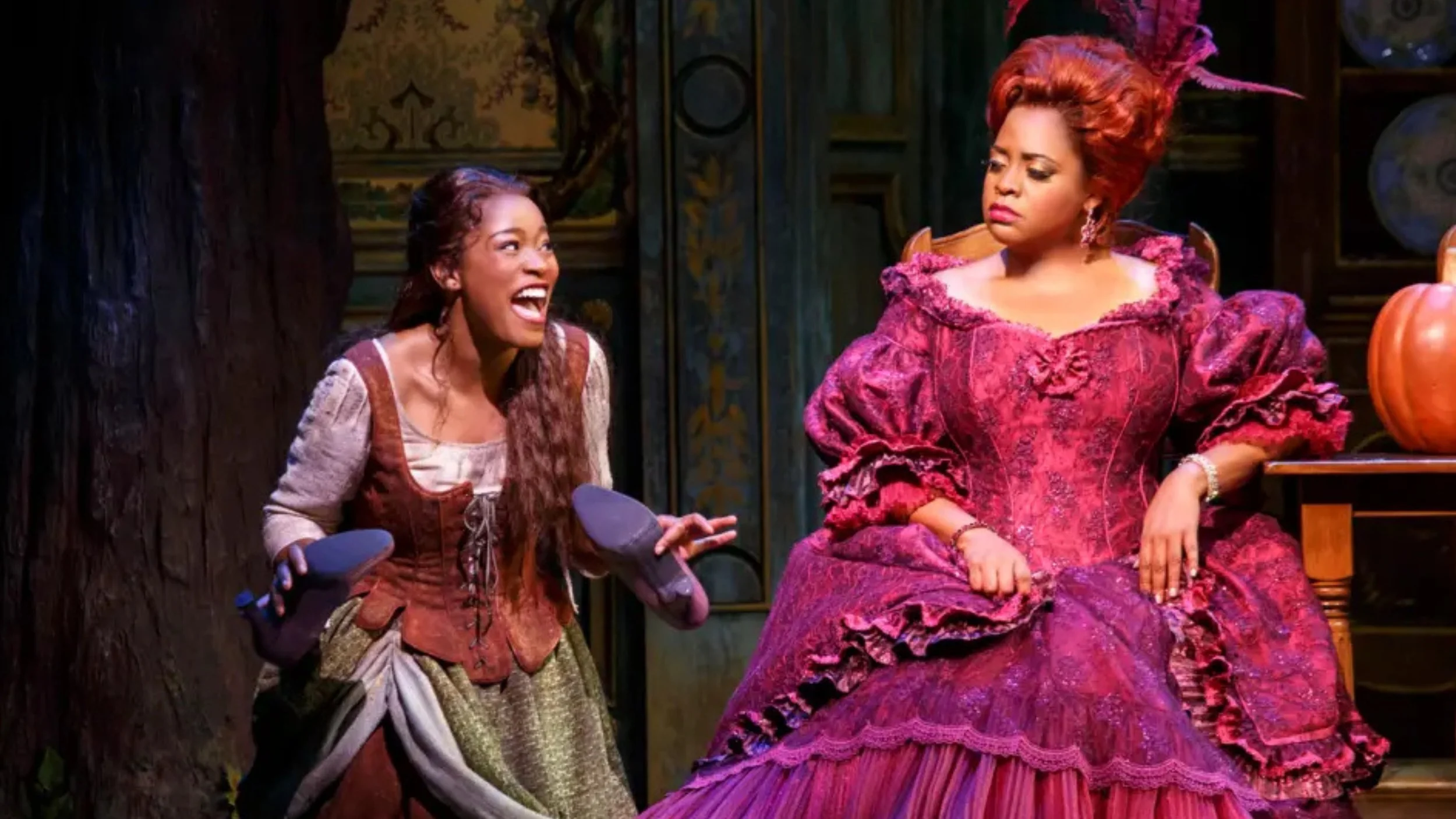Keke Palmer, in a peasant dress and holding a pair of shoes, laughs as Sherri Shepherd, in an ornate magenta gown and towering red wig with feathers, sits beside her with an unimpressed expression on a storybook stage set