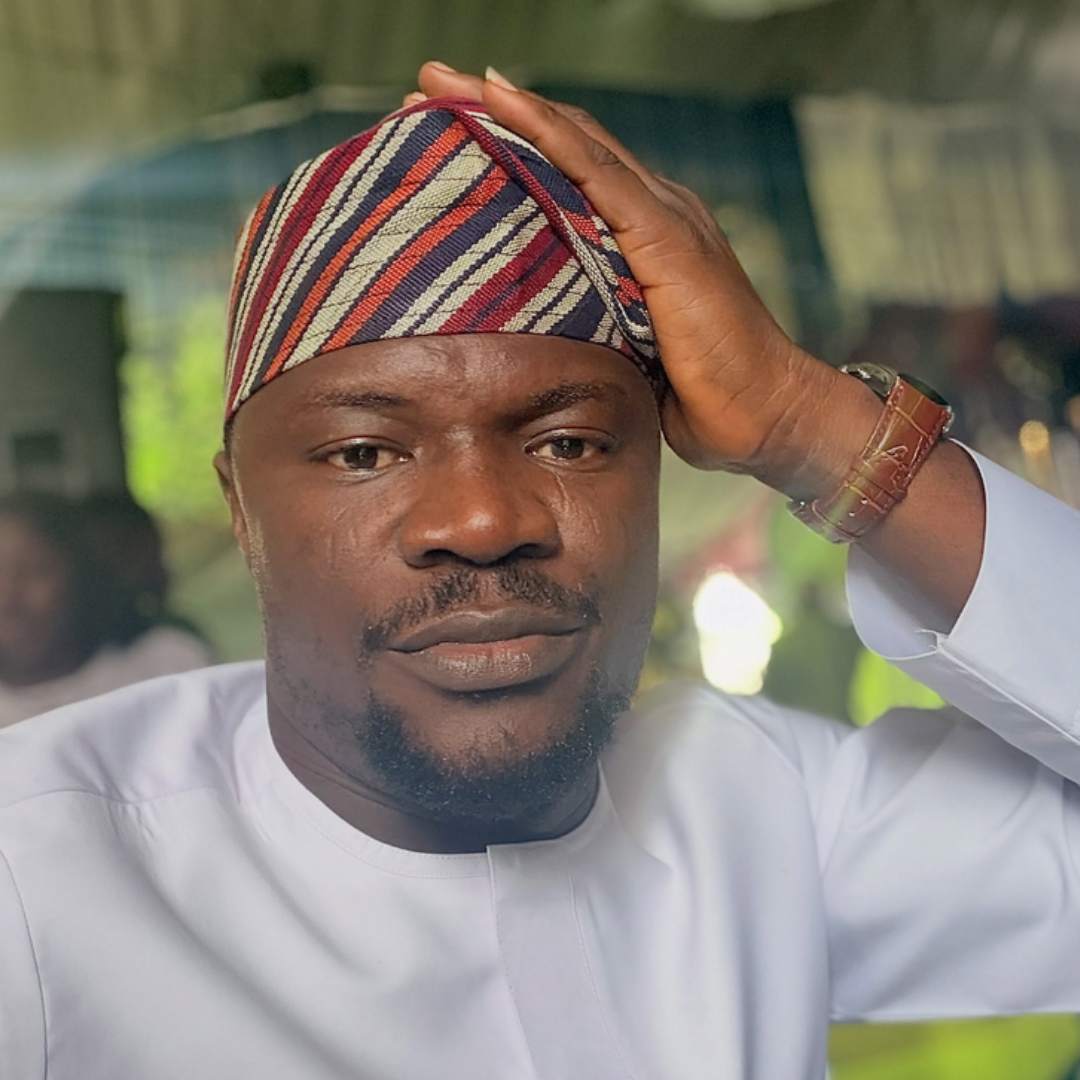 Gospel singer Toluwanisings in a white traditional outfit, one hand resting on his striped cap, with a calm, reflective expression