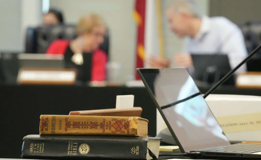 Close-up of a stack of books including a New King James Version Bible and a copy of Resurrection by Leo Tolstoy on a desk in a government meeting room, with blurred figures of board members working at laptops in the background.