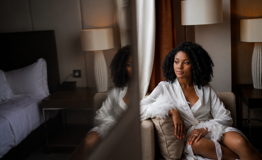 A Black woman in a white robe sitting by a window in a bedroom, reflected in a mirror beside her