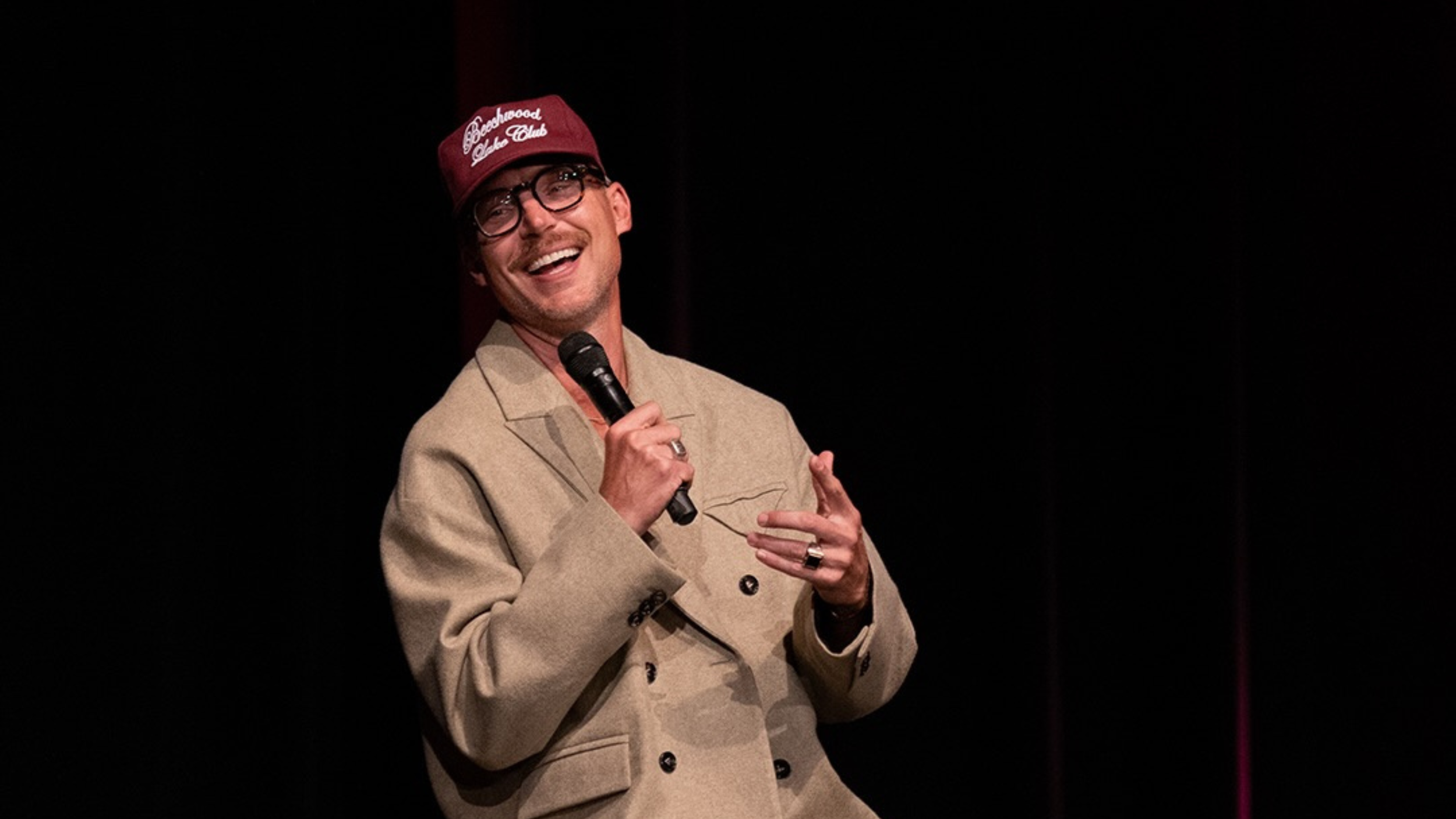 Pastor Judah Smith, smiling and wearing glasses, a maroon cap and a beige double-breasted coat, holds a microphone and gestures while speaking on a dark stage.