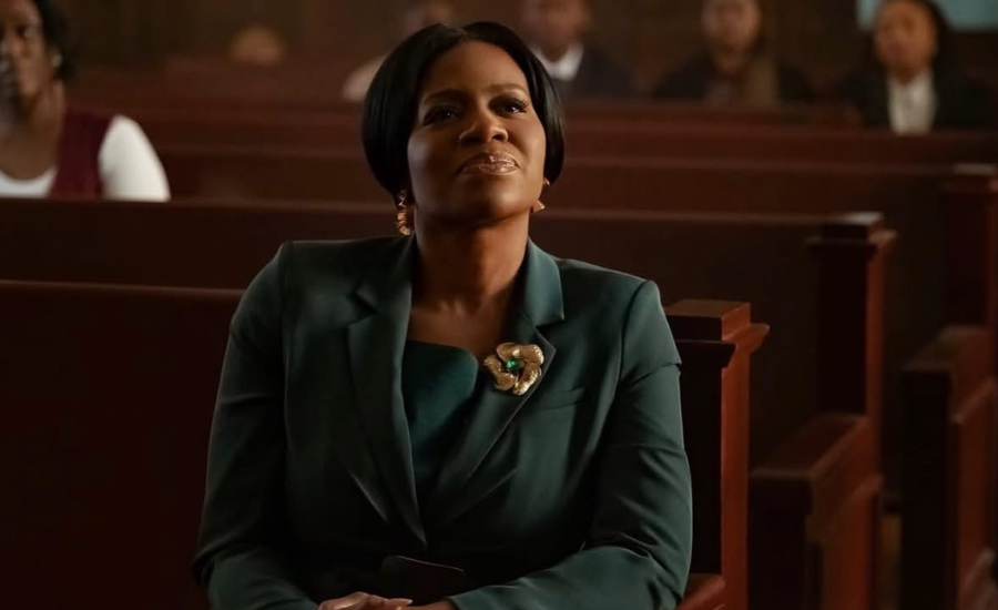 A woman in a teal suit with a gold brooch sits in a church pew, looking upward.