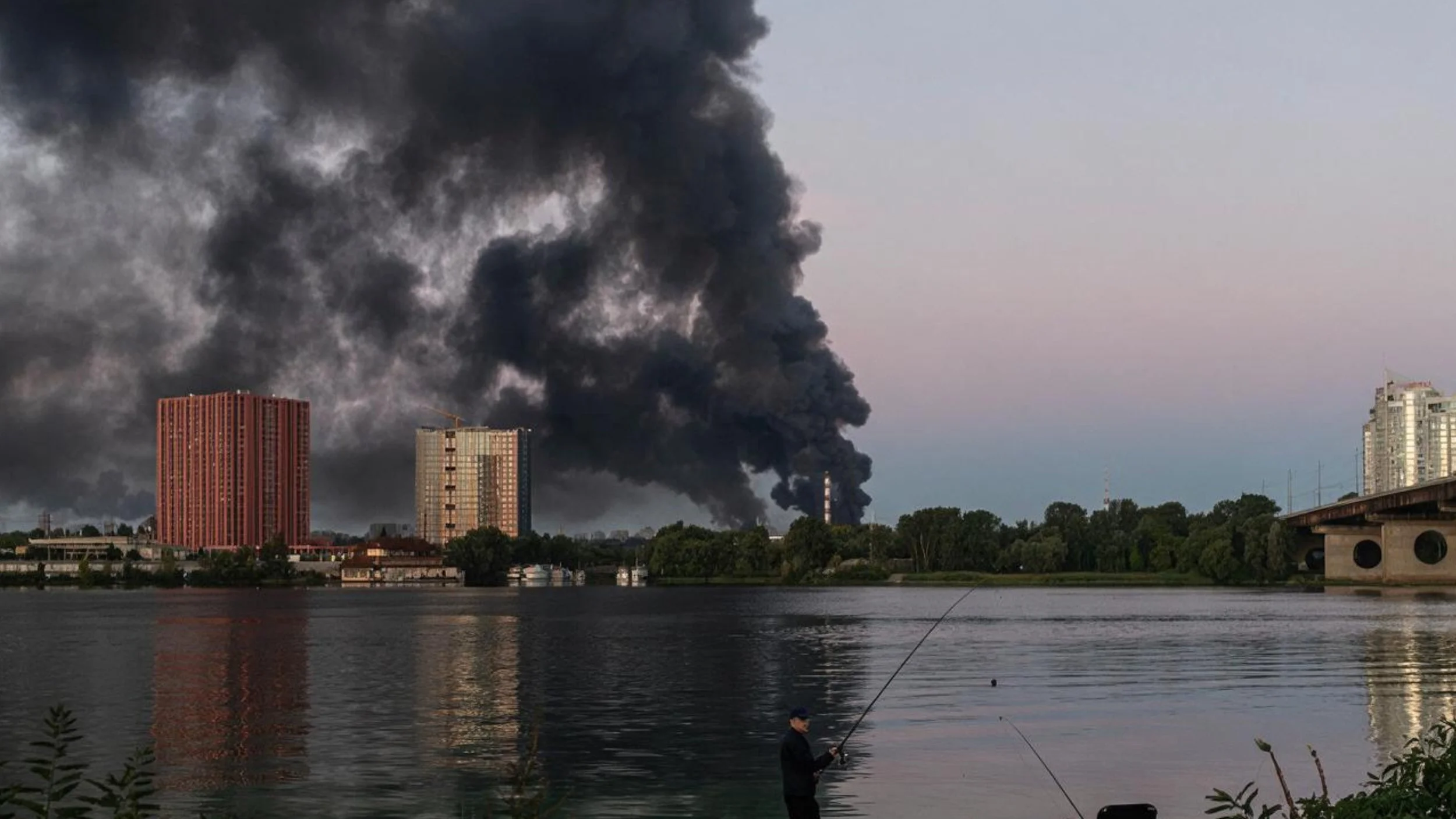 A man fishes from a riverbank in Kyiv while a massive plume of black smoke rises behind high-rise apartment buildings and trees across the water.