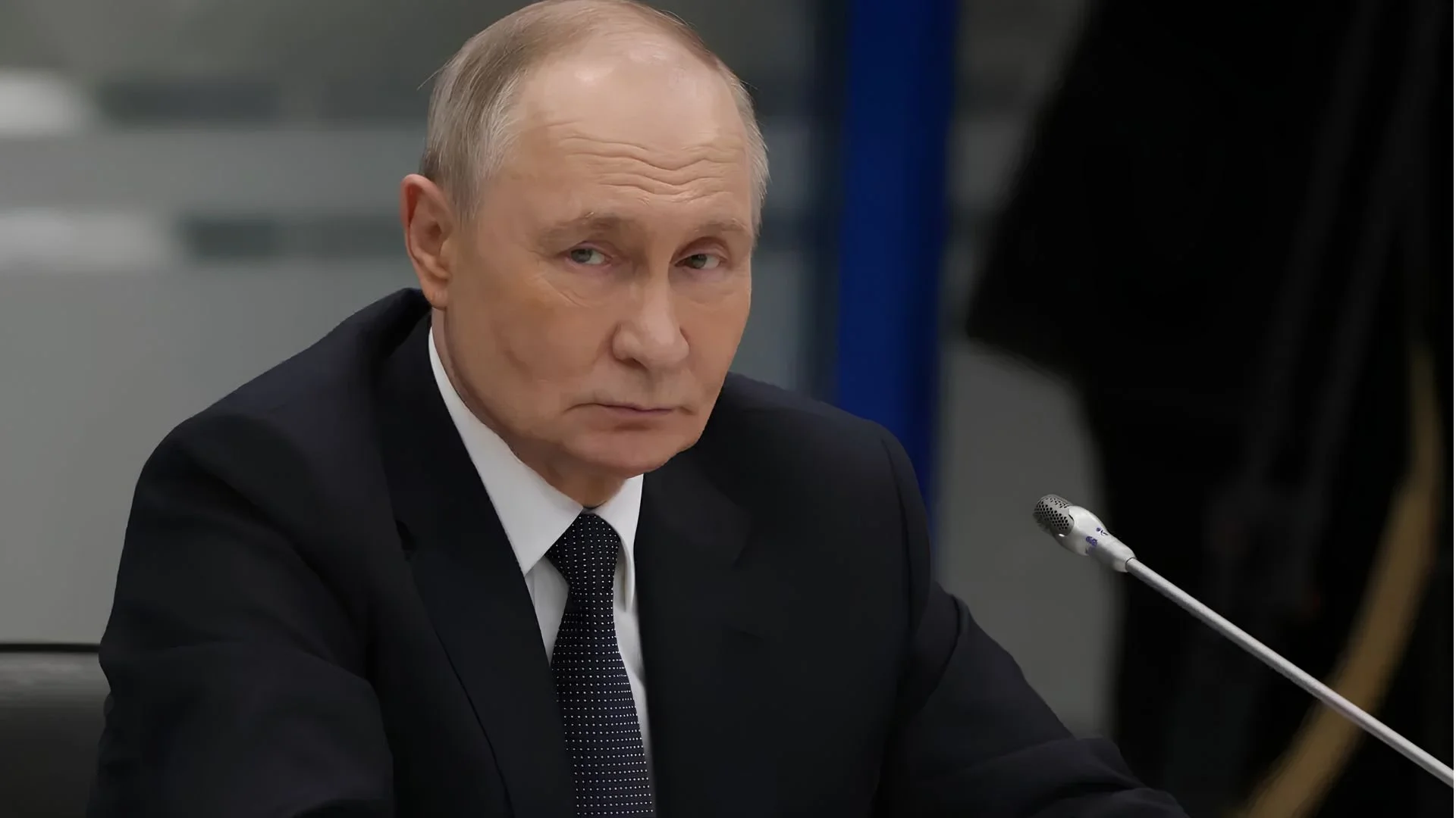 Russian President Vladimir Putin, in a dark suit and tie, sits at a microphone and looks toward the camera.
