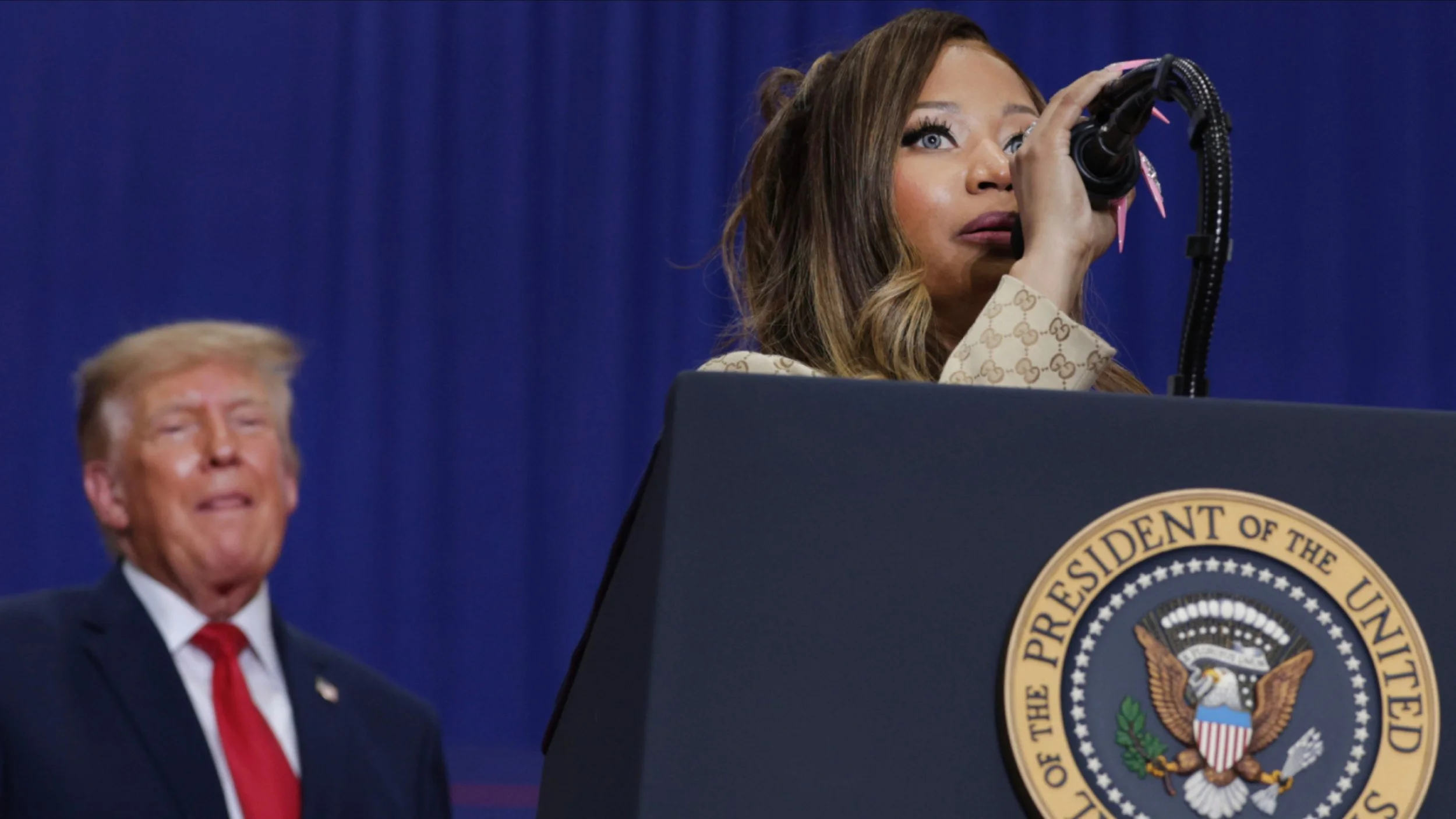 Nicki Minaj, in a tan monogrammed jacket, holds a microphone at a podium bearing the presidential seal as President Donald Trump looks on from the left.