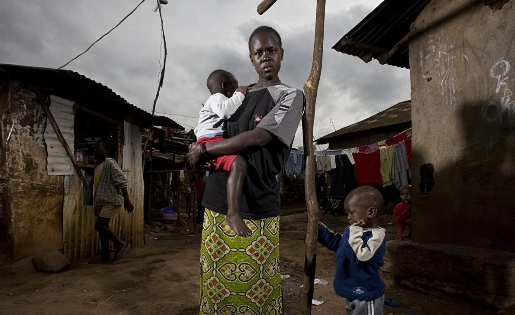 A woman in a dark T-shirt and a green patterned skirt holds a toddler on her hip in a dirt lane between tin-roofed homes, as a small boy stands beside her and laundry hangs on a line behind them under a cloudy sky.