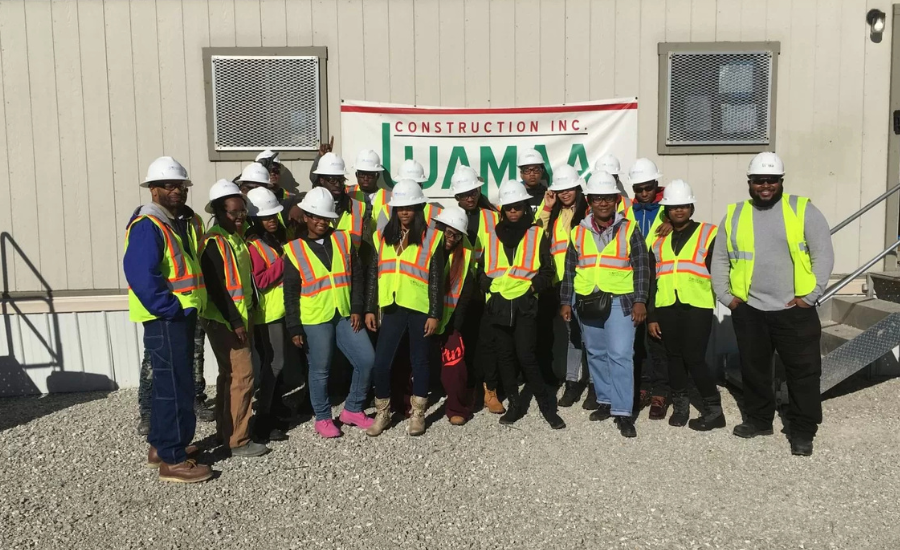 A group of UJAMAA Construction workers wearing hard hats and high-visibility safety vests pose together in front of the company's banner at a construction site
