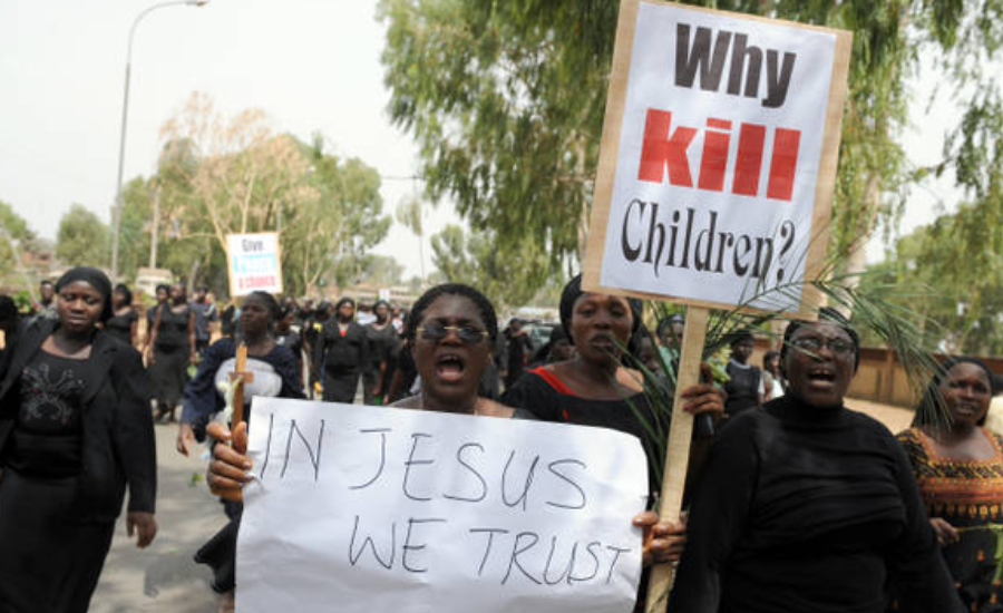 Women dressed largely in black march in protest on a Nigerian street. One woman holds a sign reading "Why Kill Children?" while another carries a handwritten sign that says, "In Jesus We Trust."