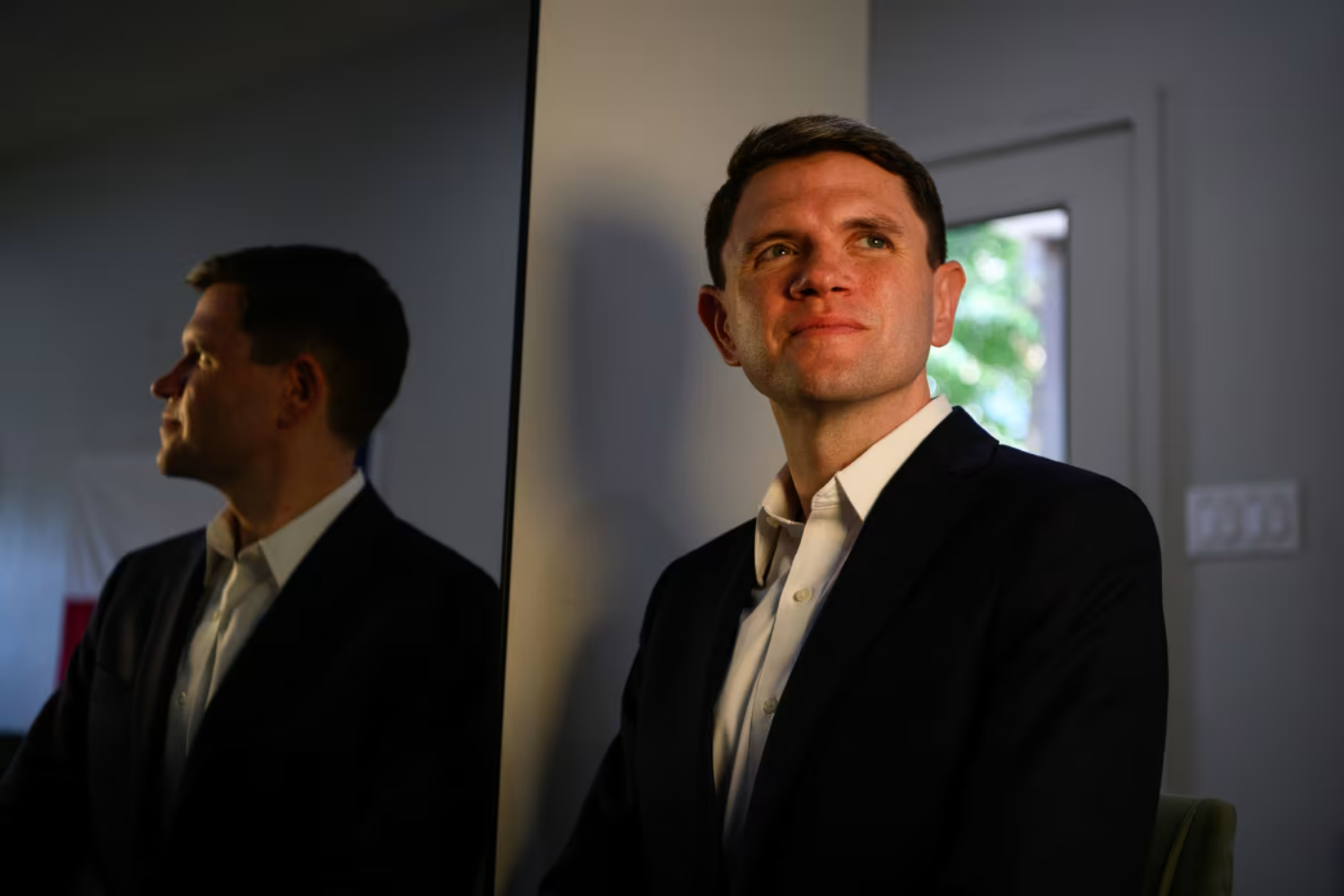 James Talarico, in a dark blazer and white open-collar shirt, looks upward in his Austin campaign office, his profile reflected in a mirror beside him.